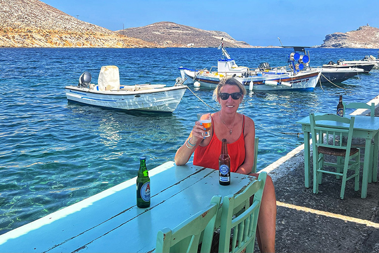 A woman in an orange top and sunglasses raising a glass of beer at a green table on the shore of a greek seaside resort with bobbing boats behind her