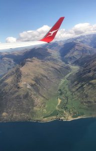 The distinctive red and white tail of a Quantas aeroplane soaring above a beautiful green mountain range and deep blue lake
