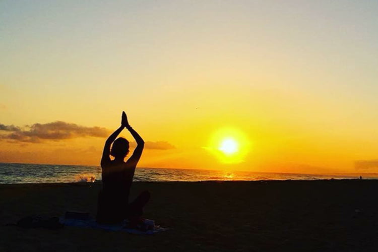 A lone silhouetted woman doing a yoga pose facing a dramatic yellow sunrise
