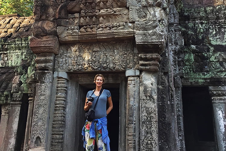 A blonde woman dressed in blue with a camera around her neck standing in front of an ancient temple in Cambodia