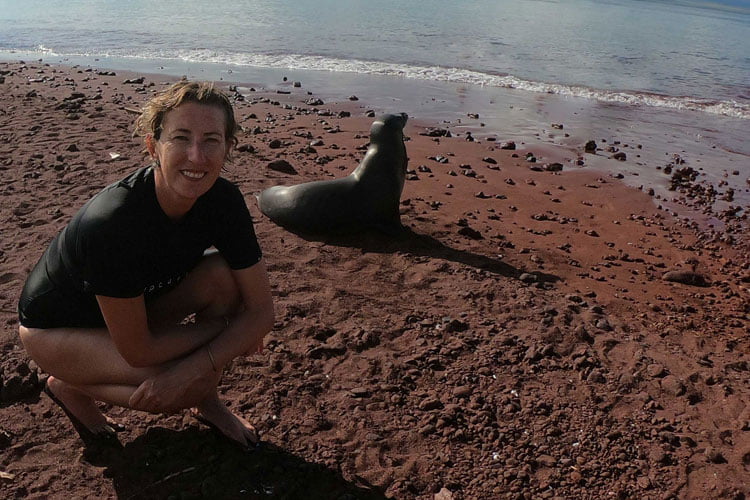A woman in a black wetsuit top crouching next to a black seal on a red sand beach by the sea