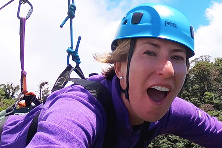 A blonde woman in purple top and blue helmet Screaming in joy while Ziplinning, in Costa Rica