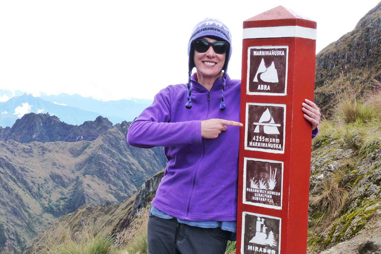 A Woman in sunglasses and purple wooly hat and fleece leaning against a red sign post with a dramatic mountainous backdrop