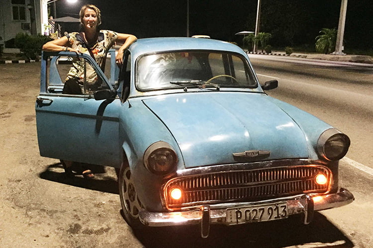 A blonde woman leaning against the one door of a blue vintage car beside a road at night with the lights on