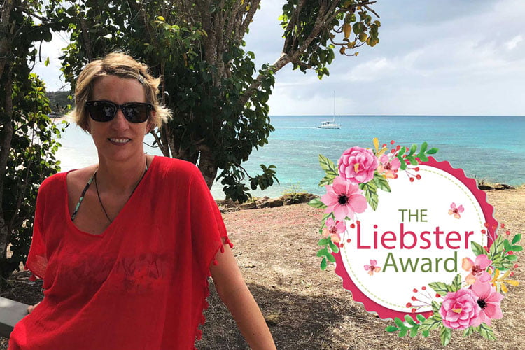 A blonde woman in sunglasses and a red top standing in front of a tree on a beach with a small boat out to see with a flowery circle overlay with the words The Liebster Award within it