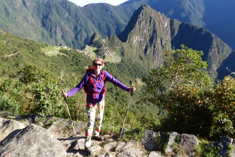 A woman in sunglasses, purple top and ski polls standing smiling with a backdrop of Machu Picchu in Peru behind