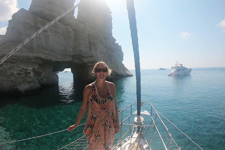 A blonde Woman standing at the end of a boat in front of a large rock with a big hole in it on a sunny day