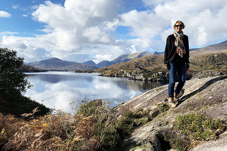 A blonde woman standing on a rock beside a mirrored lake in a remote parkland with mountainous backdrop on a cloudy day