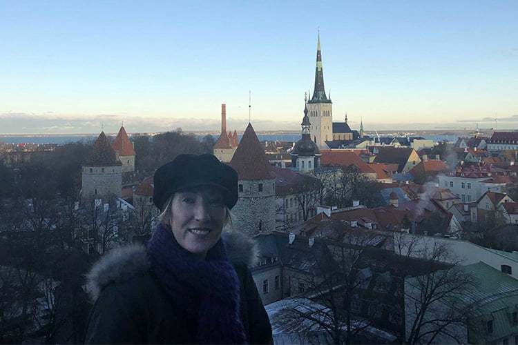 A blonde woman in a warm hat and coat with a view of Tallinn behind her
