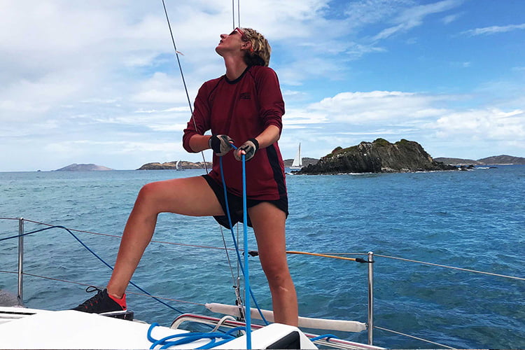 A blonde woman in a red top and sunglasses pulling on a blue rope aboard a yacht in the caribbean