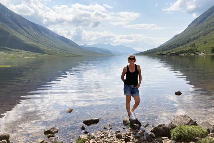 A Woman in a black vest and sunglasses standing beside a calm loch in Scotland with mountains each side of the water