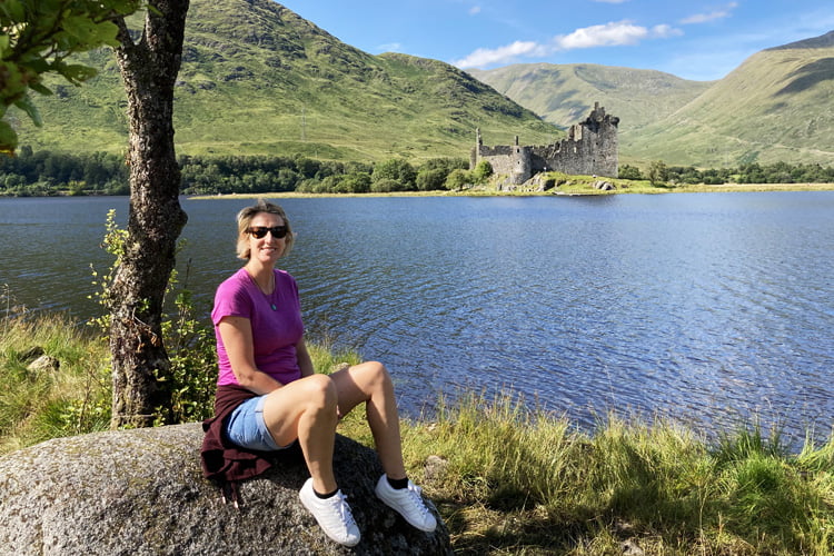 A Woman in a pink top sitting on beside a calm loch in Scotland with a ruined castle and mountains in the far distance