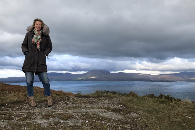 A Woman in a green coat with fur hood standing on a remote hillside overlooking a flat overcast seascape
