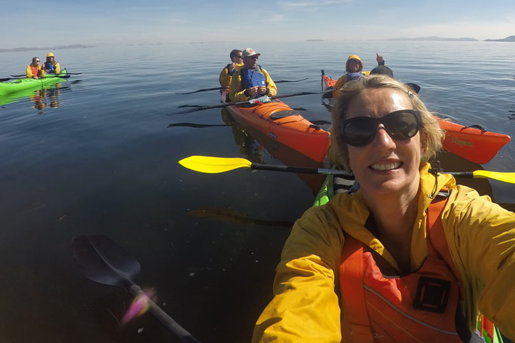 A blonde woman in sunglasses and yellow waterproof taking a selfie from a green canoe on a flat lake with other duos behind in orange and green canoes