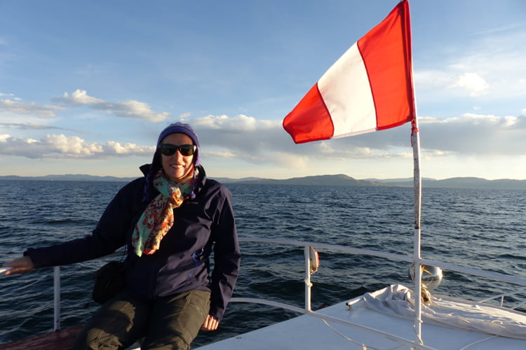 A Woman in sunglasses blue coat and jacket sitting on a boat on a choppy sea next to a re and white Peruvian flag