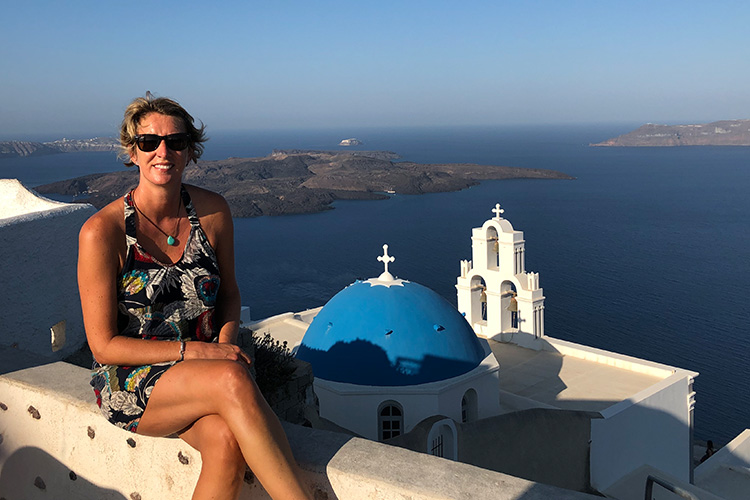 A Woman in sunglasses and summer dress sitting high on a hill overlooking a blue domed Greek Church and a flat blue sea and island setting