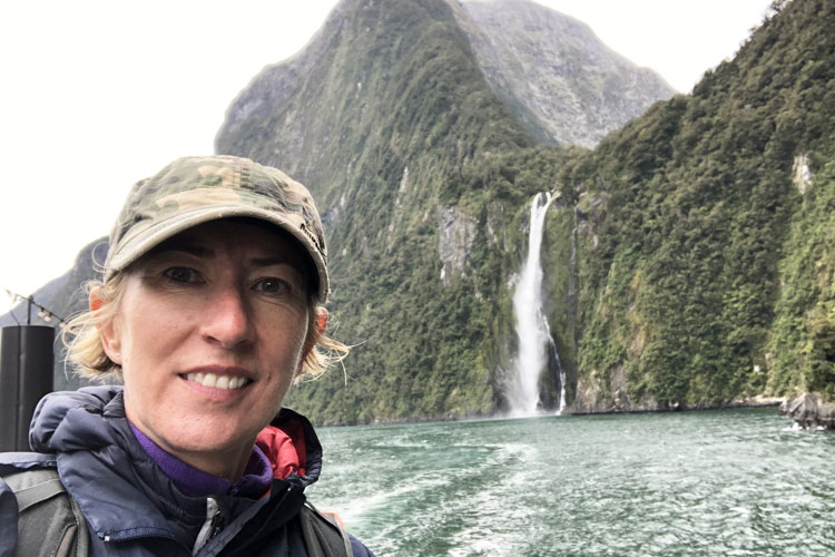 A Woman in a camouflage Cap standing in front of a dramatic mountain waterfall