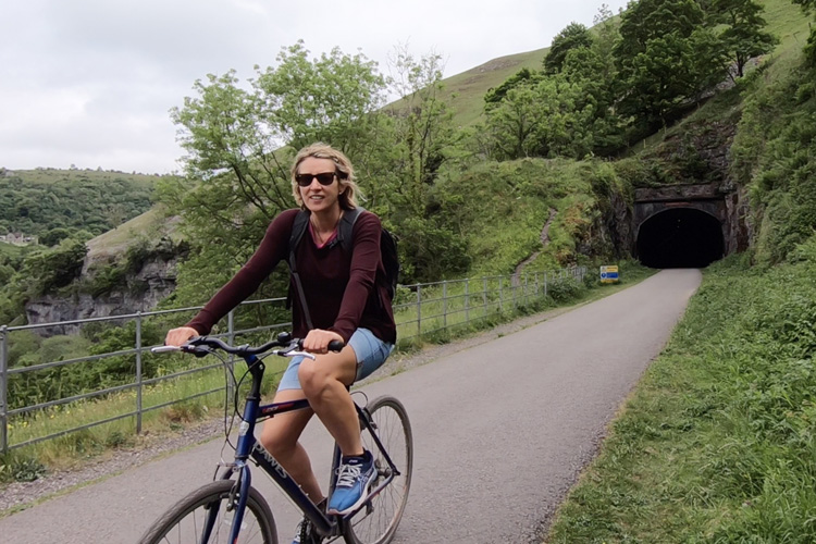 A blonde woman in sunglasses and shorts riding a bike along a tarmac path leading away from a circular tunnel on a green hillside
