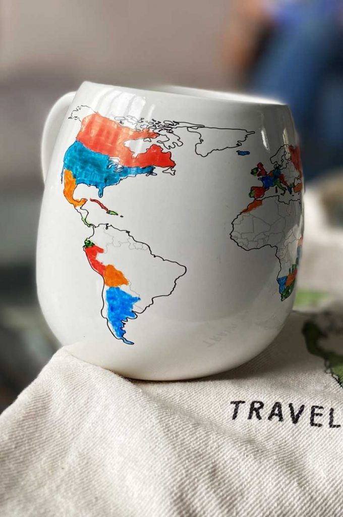 A white coffee mug with a handrawn world map that you can colour yourself sitting on a tote bag on a table