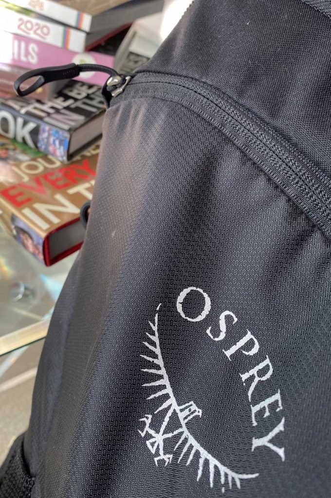 A close up of a grey rucksack with the word osprey printed in white on it, in front of a pile of travel books