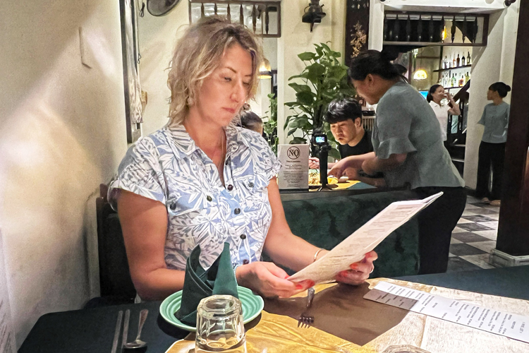 A Woman in a light blue top reading a menu sitting alone at a dinner table in a restaurant