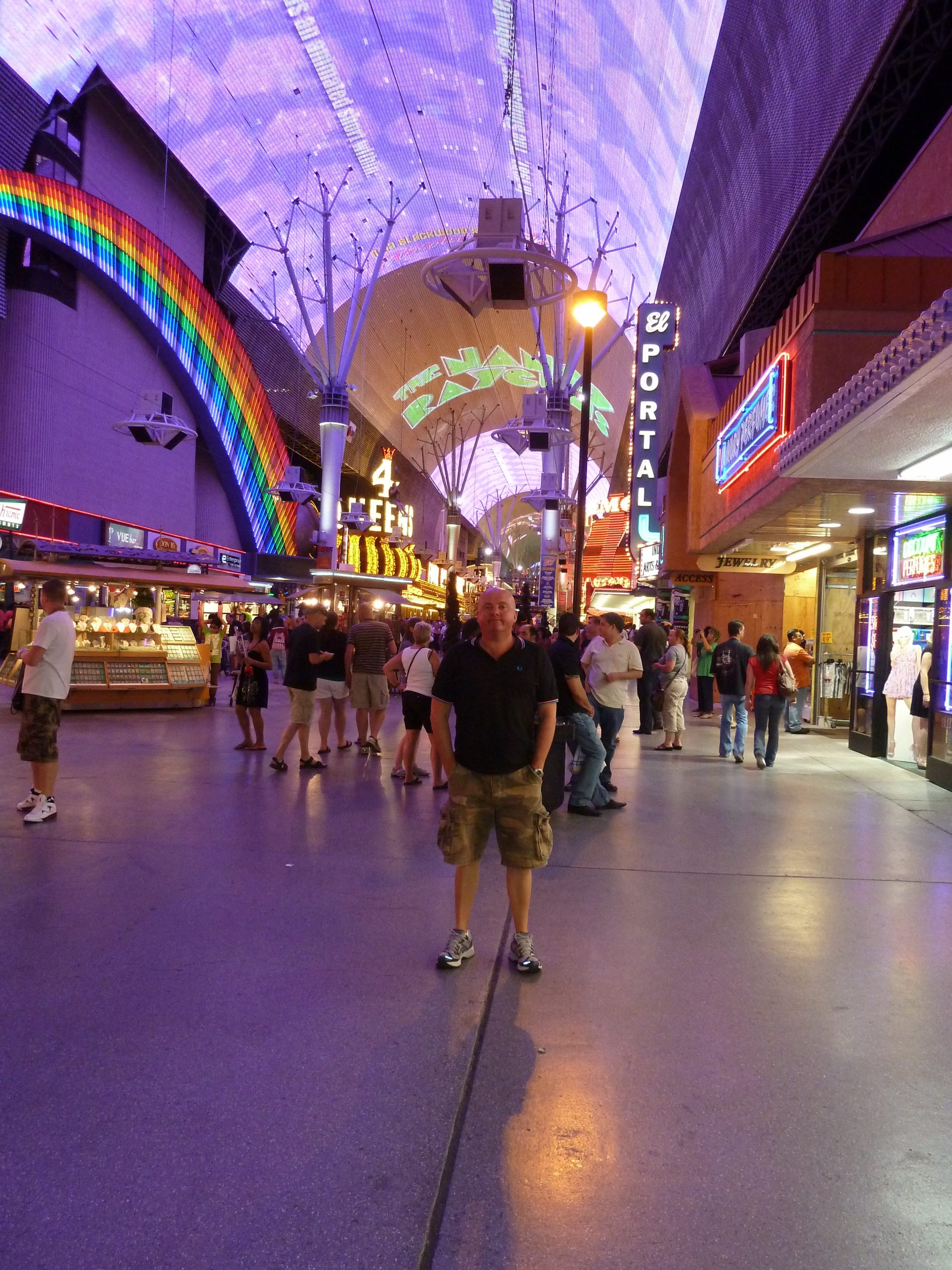 A man in shorts standing in a brightly lit shopping parade in the evening