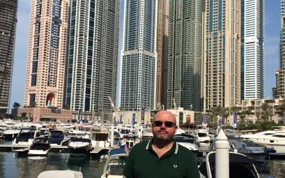 A man in sunglasses and black t-shirt standing in front of huge skyscrapers in Dubai
