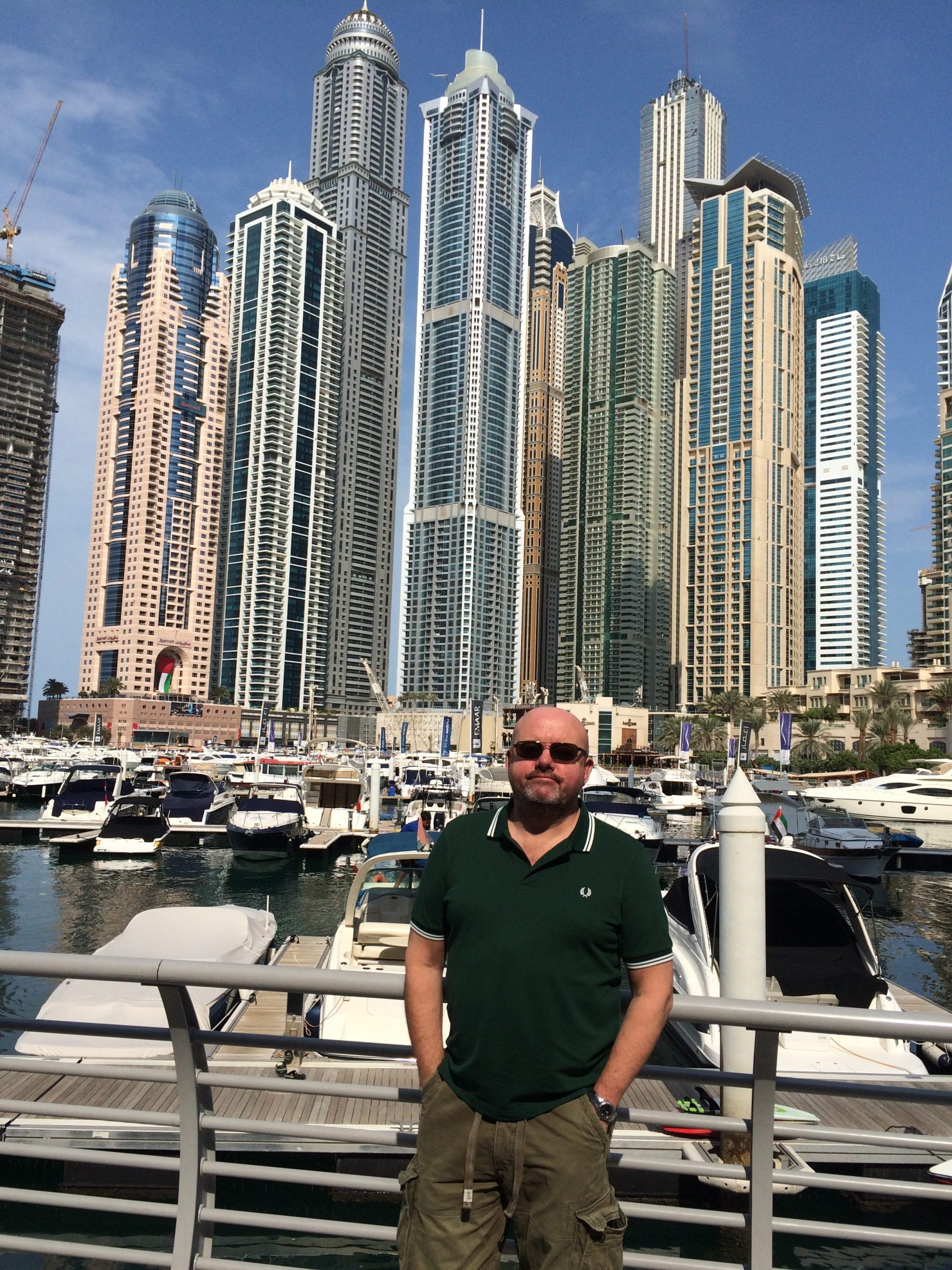 A man in sunglasses and black t-shirt standing in front of huge skyscrapers in Dubai
