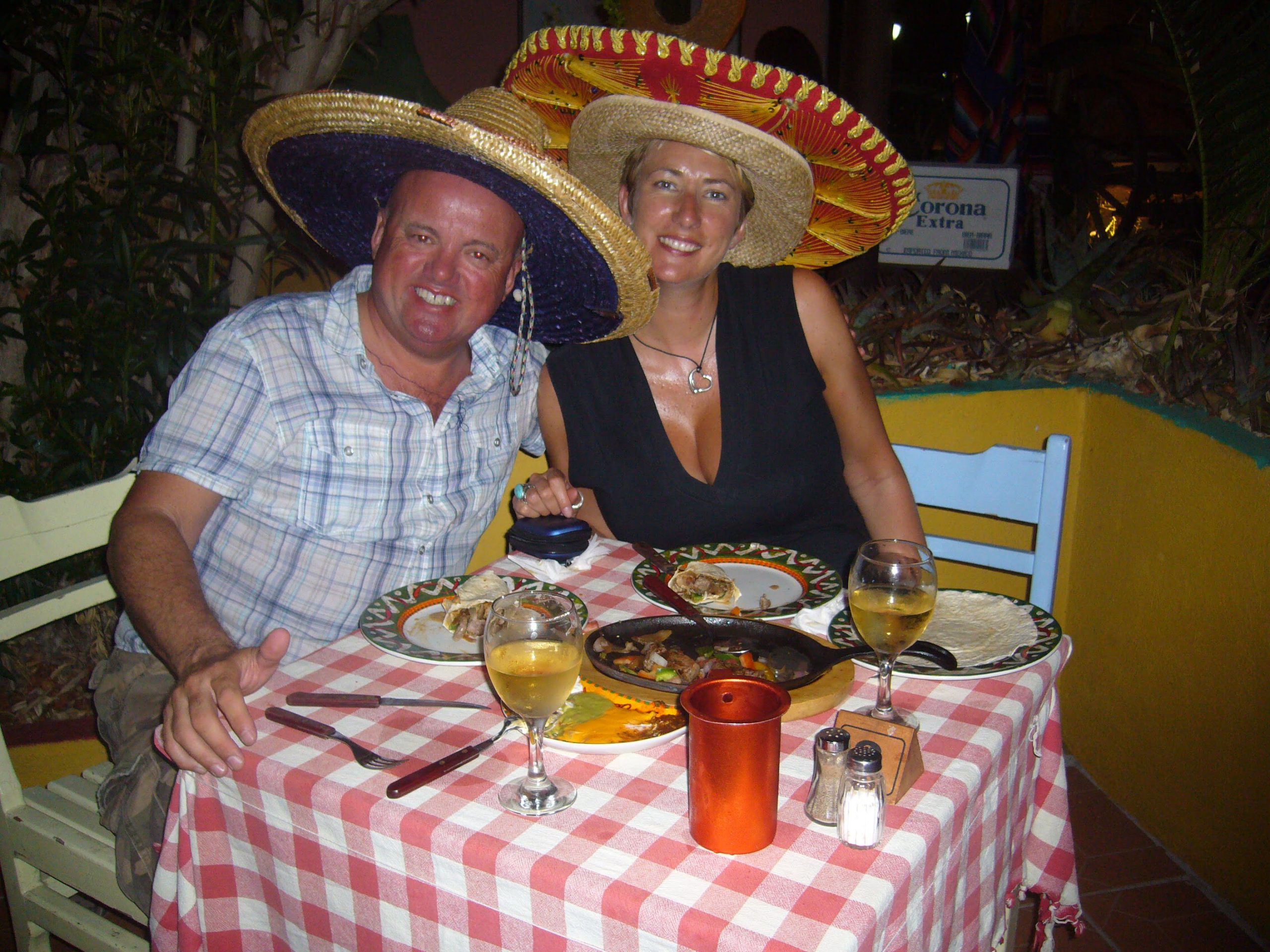 A smiling couple sitting at a dinner table covered in a checked cloth wearing Sumbreros