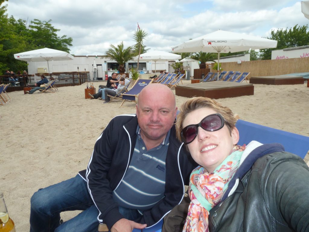A picture of a bald man in a striped t-shirt having a selfie with a blonde woman in sunglasses sitting having a beer on a deck chair on sand