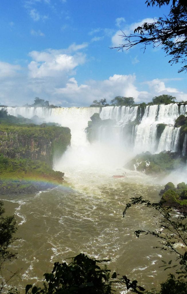 Raging white water falls in a sheer wall of steam causing a rainbow to rise from the brown turbulent waters of Iguazu falls