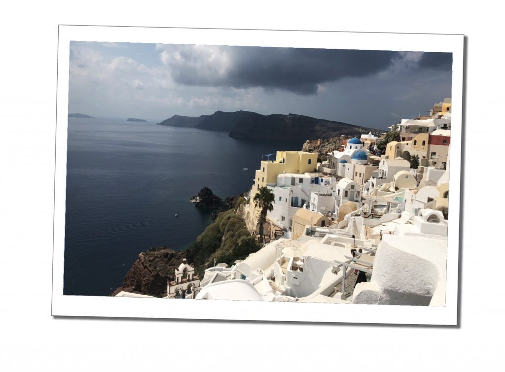 Slate grey skies with a hint of sunlight over the beautiful panorama of Santorini's whitewashed buildings that cling to the hillside overlooking the bay.