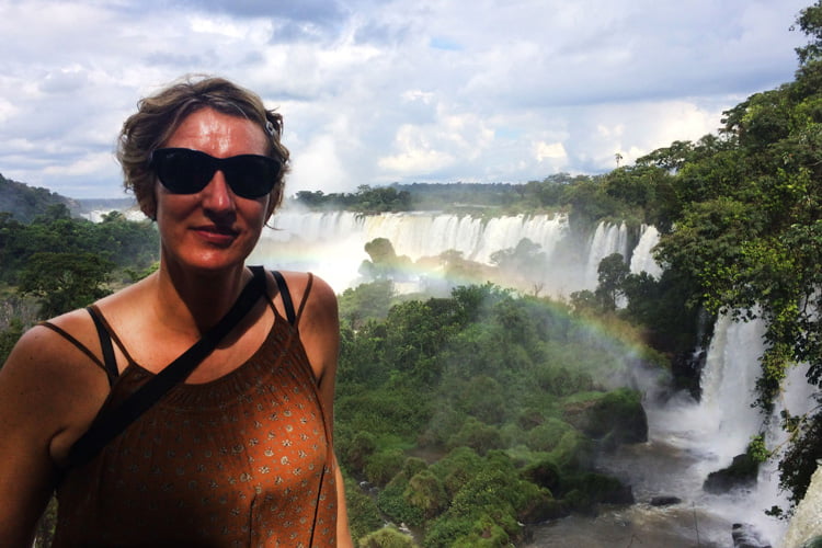 A blonde woman in sunglasses and brown top stands high above a wall of cascading waterfalls punctuated by a misty rainbow