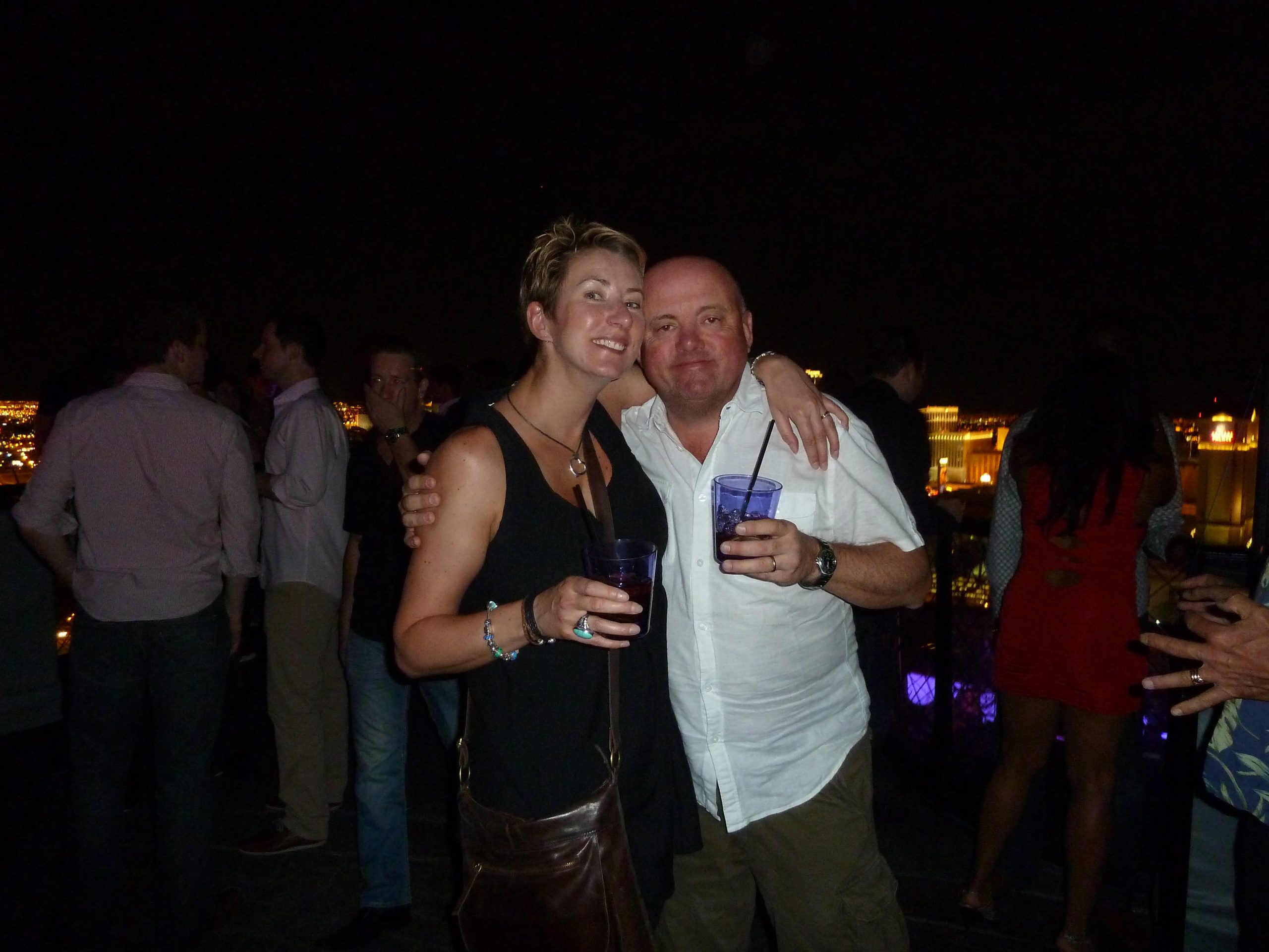 A blonde woman and a man standing in a bar holding cocktails surrounded by people and flickering lights