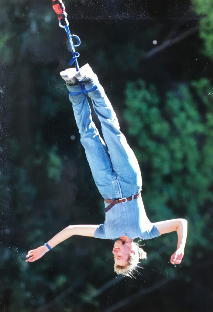 A blonde woman upside down while Bungee Jumping from a bridge in Queenstown, New Zealand wearing jeans and a blue top