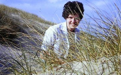 A woman with black hair crouching behind tall grass in sand dunes