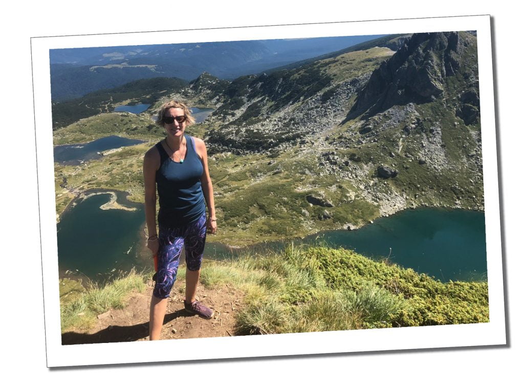 A Woman in dark glasses and sports trousers in front of a kidney shaped lake high on a green hill on a cloudless sunny day