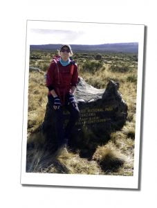 A woman in a red top, cap and sunglasses sits resting on a rock with an inscription on it on the tundra of Kilimanjaro
