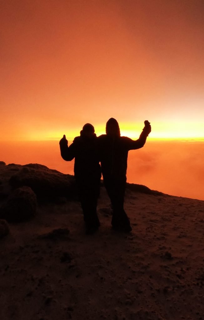 Two Women standing in silhouette at sunrise at the top of a frozen snowy mountain