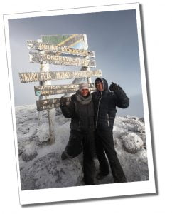 Two Women in a warm blue coat with her arms spread wide standing in front of a large sign post at the top of a snow covered mountain