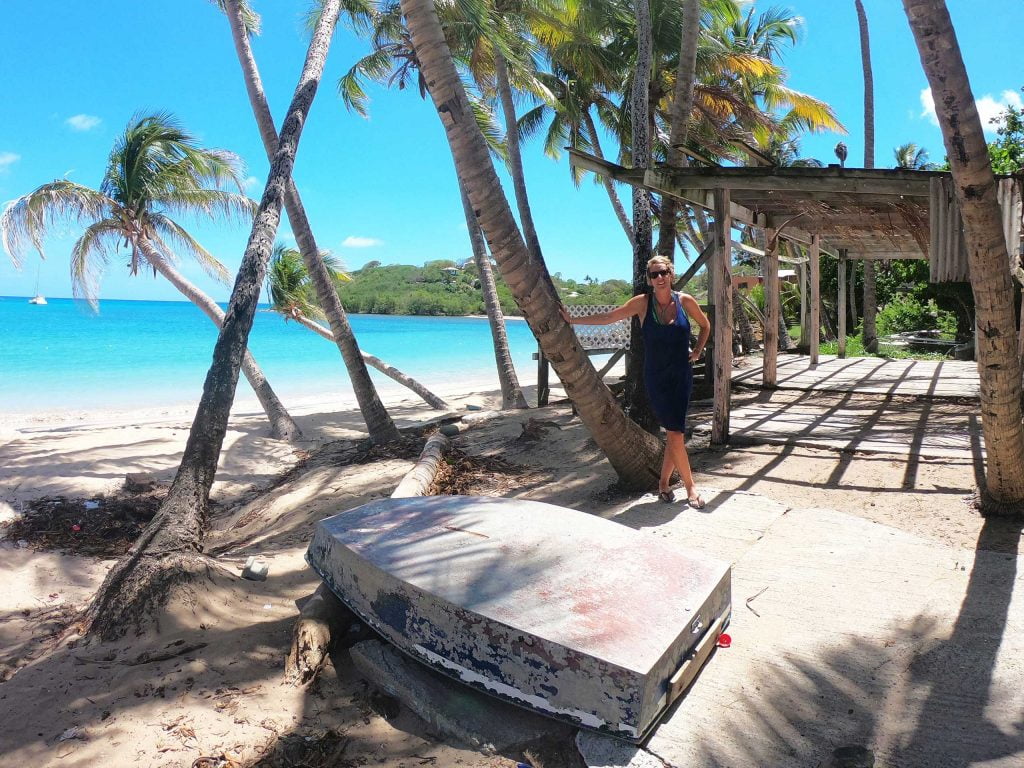 A blonde woman in a black dress and sunglasses standing leaning against a palm tree on a caribbean beach with an upturned boat in the foreground