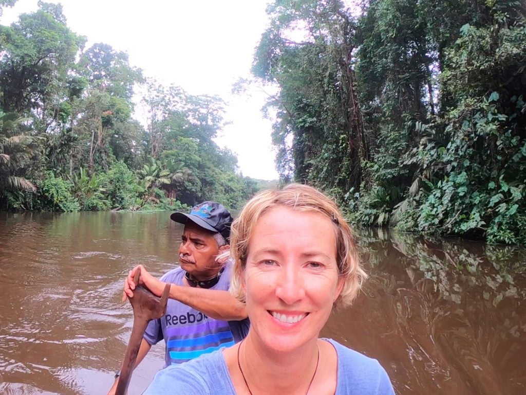 A blonde woman sitting in a canoe with a man rowing behind her on a brown river in a tropical setting
