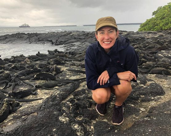 A blonde woman in a cap crouching on a rocky beach in front of a group of marine iguana