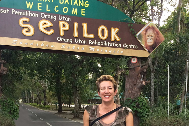 A blonde woman standing under a big sign that say Sepilok