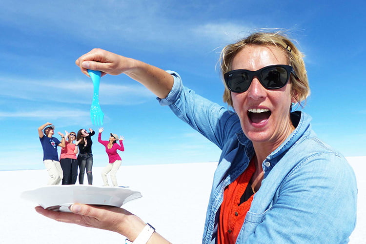 A blonde woman in denim jacket and sunglasses by using an optical illusion is pretending to put a plastic for into 4 people standing on a plate