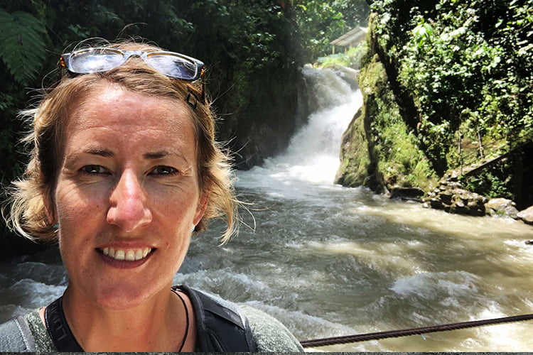 A blonde woman with sunglasses on her head standing in front of a raging white water stream