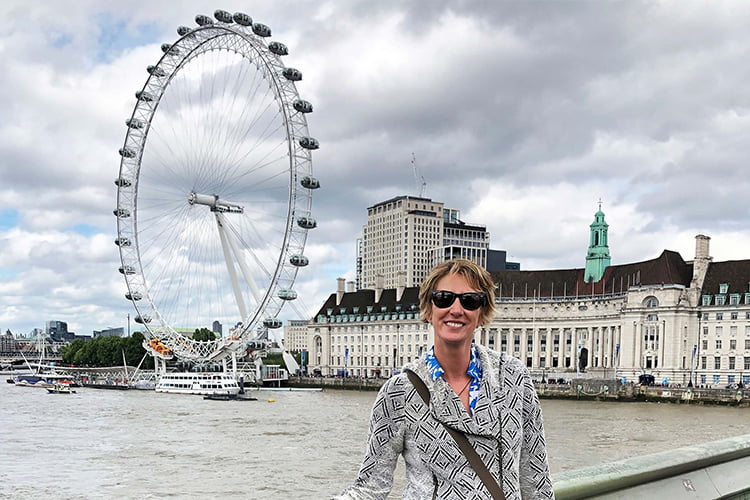 A blonde woman in sunglasses and cardigan standing on a london bridge overlooking the river thames and london eye