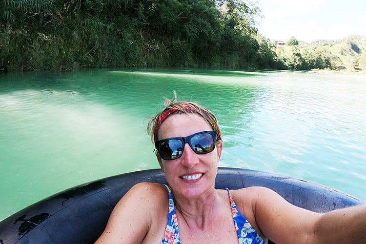 A blonde woman in sunglasses sitting in a black rubber tyre floating along a green river