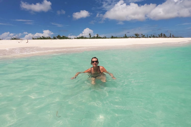 A blonde woman in sunglasses floating in a clear Caribbean sea by a deserted beach