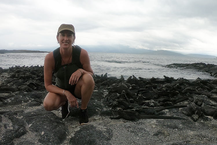 A blonde woman in a cap crouching on a rocky beach in front of a group of marine iguana